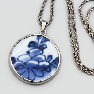 May include: A silver-toned necklace featuring a round pendant with a blue and white floral design. The pendant is framed in silver and hangs from a matching silver chain. The necklace is displayed against a light gray background.