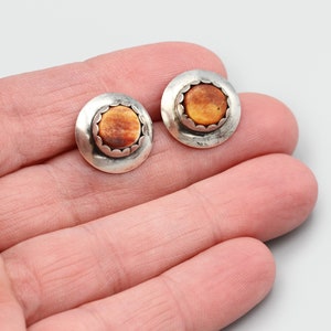 Puede incluir: Un par de pendientes de botón redondos. Cada pendiente presenta un borde plateado festoneado que rodea una piedra central, circular, de color naranja-marrón. Los pendientes se muestran sobre un fondo claro.