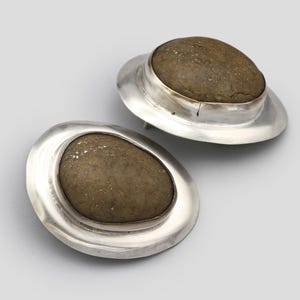May include: A pair of stud earrings featuring smooth, oval-shaped brown stones set within polished silver bezels. Each earring has a wide, flat, circular silver frame surrounding the central stone, creating a layered, metallic appearance.