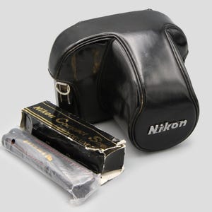 May include: A black Nikon camera case with the Nikon logo in silver. Beside it is a black box with gold lettering that reads "Nikon Compact Series Deluxe". A clear plastic package is next to the box.