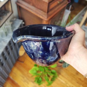 Puede incluir: Un cuenco de cerámica azul oscuro hecho a mano con un acabado brillante. El cuenco tiene una forma única con un pico vertedor y está decorado con un patrón moteado. El cuenco es sostenido por una persona.