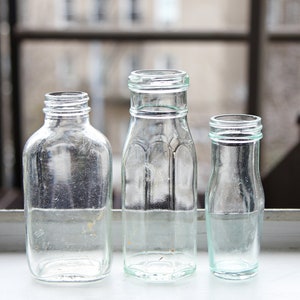 Puede incluir: Tres botellas de vidrio transparente de diferentes formas y tamaños están dispuestas en un alféizar. Las botellas están vacías y tienen una estética vintage. El fondo está borroso, lo que sugiere un entorno al aire libre.