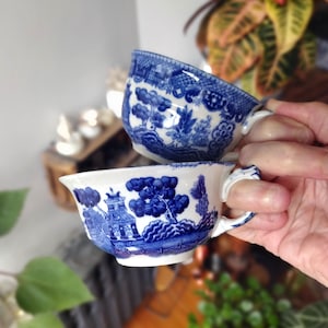 May include: Two blue and white porcelain teacups with a classic Willow pattern. The cups are held in a hand, showcasing their detailed landscape design and curved handles. The teacups are a classic design.