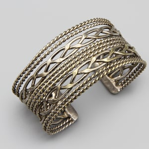 May include: Silver cuff bracelet with an intricate openwork design. The bracelet features interwoven patterns and twisted rope-like edges. The open design allows for flexibility and a comfortable fit. A statement piece for everyday wear.