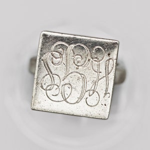 Puede incluir: Anillo de plata con una cara cuadrada grabada con iniciales ornamentadas e entrelazadas. El anillo tiene una banda sencilla y una apariencia ligeramente envejecida, lo que sugiere que es vintage. El diseño es clásico y elegante. La cara mide aproximadamente 1,5 cm x 1,5 cm.
