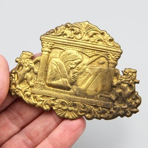 Puede incluir: Una placa de metal dorado con un diseño ornamentado que presenta una figura arrodillada y escribiendo. La placa está decorada con intrincados detalles florales y arquitectónicos.