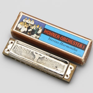 May include: A silver Hohner Vereins-Harmonika Orchester I harmonica in its original box. The box has a blue and white label with a picture of a group of people playing music. The harmonica has the words "Hohner Vereins Harmonika Orchester I" printed on it.