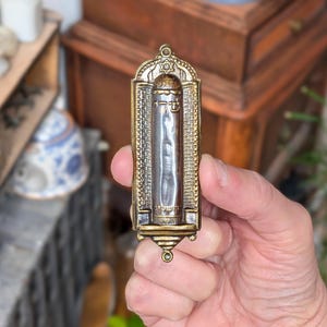 May include: A vertical, rectangular mezuzah case made of antique-toned brass. The central cylindrical section features Hebrew lettering and a Star of David at the top. The frame has a textured, brick-like pattern on both sides and a small loop at the top.