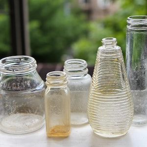Puede incluir: Cinco botellas de vidrio transparente de diferentes formas y tamaños. Las botellas están vacías y tienen un aspecto vintage. Algunas de las botellas tienen una superficie texturizada.
