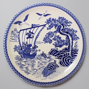 May include: A round, white ceramic plate with a blue and white Asian-inspired design. The plate features a sailing ship, birds, and a detailed border. The design includes a tree, waves, and a turtle.