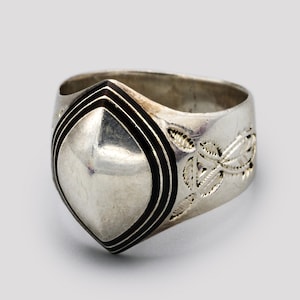 Può includere: Anello d'argento con un design centrale a forma di scudo bordato di nero. La fascia dell'anello presenta un motivo floreale. L'anello è realizzato in un materiale metallico lucido.