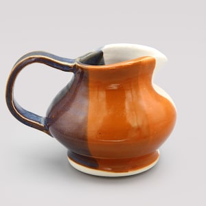 May include: A ceramic creamer pitcher featuring a rounded body with a glossy finish. The exterior is divided into sections of deep navy blue, warm orange, and white. It includes a curved handle on the side and a small pouring spout at the top rim.