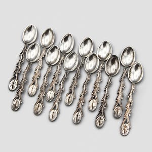 May include: A collection of eleven silver spoons with ornate, floral-inspired handles. Each spoon features a rounded bowl and a detailed, leaf-like design extending down the handle. The spoons are arranged on a light gray background.