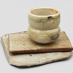 May include: A handmade ceramic cup and two matching plates. The cup is a light beige with brown and black markings. The plates are rectangular with a similar color scheme and a dark brown edge. The set has a rustic, artisanal feel.