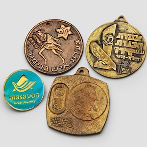 Può includere: Quattro spille in metallo vintage con diversi design. Una spilla è blu con un design a onde e il testo "masa'von Israel Journey". Un'altra spilla è in bronzo con una donna che trasporta un oggetto grande e il testo "20 שנה לעיר" in ebraico. La terza spilla è in bronzo con un design di una barca e il testo "עשר שנים לתנועת " in ebraico. La quarta spilla è in bronzo con un design di una persona e il testo "HERE" in inglese.