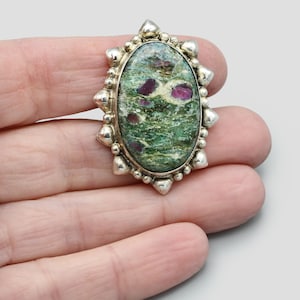 May include: An oval pendant with a silver-coloured frame. The pendant's centre features a green stone with red and white inclusions. The frame has a decorative, studded design around the edge. The pendant is held in a hand.