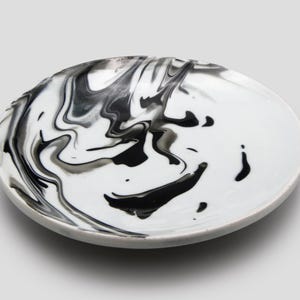May include: A round, white ceramic plate with a marbled black and gray design. The plate's surface features swirling patterns of black and gray, creating a unique, artistic aesthetic. The plate has a simple, elegant design.