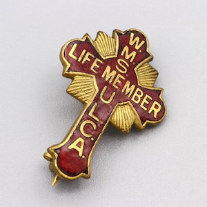 May include: A vintage gold-coloured metal pin in the shape of a cross. The pin features a red enamel centre with the words "LIFE MEMBER" and other text. The pin has a pin back for attachment.