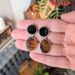 May include: A pair of silver-toned earrings. Each earring features a black, circular top and an oval-shaped, amber-colored stone with brown marbling. The earrings are held in a hand against a background of green and orange foliage.