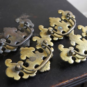 May include: A collection of five antique brass drawer pulls. Each pull features a decorative, ornate design with a curved handle. The brass has a warm, aged patina, suggesting vintage or reclaimed hardware for furniture restoration.