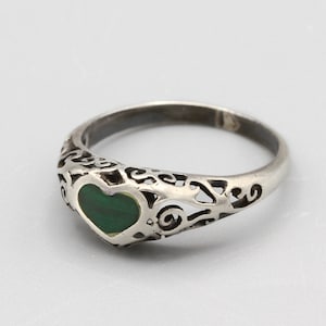 May include: A silver ring with a heart-shaped green stone at the center. The band features an openwork design with swirling patterns. The ring is set against a plain, light background.