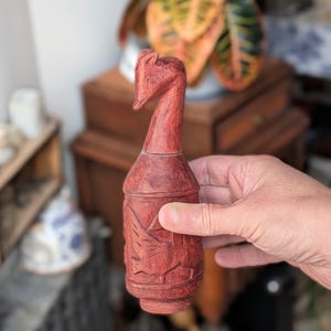 May include: A hand-carved, reddish-brown wooden bottle stopper. The stopper features a horse head design on top and carved relief details around the body. The bottle stopper is held in a person's hand.