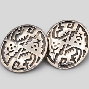 May include: Two silver-toned oval earrings with an engraved design. Each earring features a symmetrical pattern with geometric shapes and a smiling face. The earrings are displayed on a light gray background.