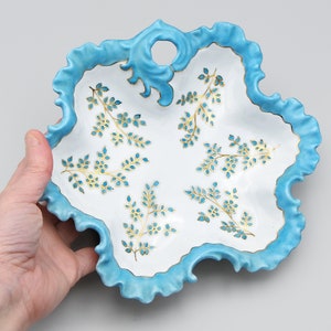 May include: A white ceramic serving dish with a light blue ruffled edge and a decorative floral pattern. The dish has a unique shape and a small handle. The floral pattern is in gold and light blue.