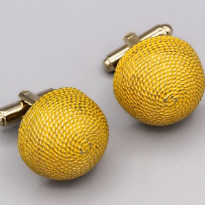 Può includere: Un paio di gemelli color oro. Ogni gemello presenta un design sferico strutturato. Il metallo ha una finitura lucida. I gemelli sono esposti su uno sfondo grigio chiaro. Accessori per abbigliamento formale.