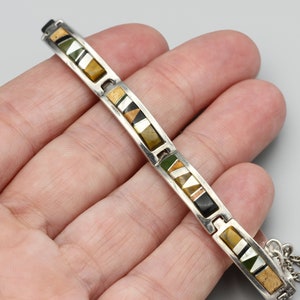 May include: Silver-toned bracelet with rectangular links. Each link features a mosaic of earth-toned geometric shapes in shades of brown, green, black, and white. The bracelet is held in a hand against a gray background.