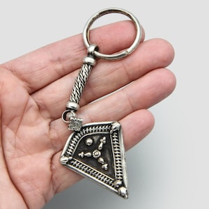 Puede incluir: Llavero de plata con un amuleto triangular decorativo. El amuleto tiene un diseño geométrico con pequeños puntos en relieve y una estrella central de tres puntas.