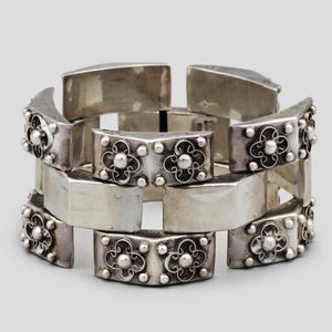 Puede incluir: Un brazalete plateado con un diseño geométrico. El brazalete está compuesto por eslabones rectangulares y trapezoidales. Cada eslabón trapezoidal presenta un diseño floral y pequeños detalles redondos.