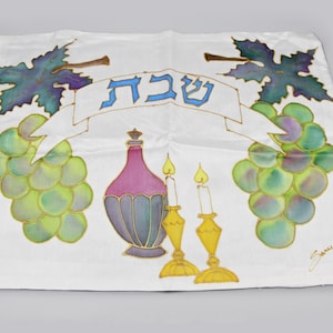 Peut inclure: Une housse en tissu blanc pour challah ornée d'un motif peint à la main représentant deux grappes de raisins verts et violets, une carafe à vin violette et deux bougeoirs jaunes avec des flammes. Le mot hébreu Shabbat est écrit en bleu.