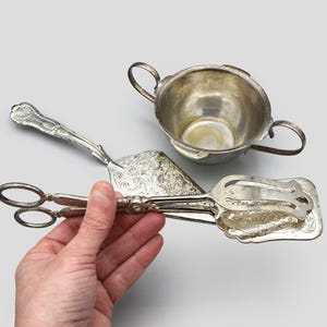 May include: Silver serving set including a sugar bowl with handles and a pair of tongs. The tongs are held by a hand. The set has ornate floral detailing. The items are made of silver.