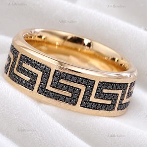 May include: A gold band ring featuring a continuous black stone inlay arranged in a geometric Greek key pattern. The ring has a polished gold finish on the edges and interior, with the black stones set flush within the central band surface.