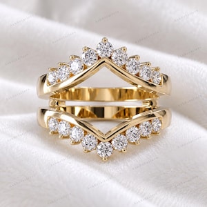 Puede incluir: Un protector de anillo de oro que presenta dos bandas en forma de cheurón conectadas en los lados. Cada banda está engastada con una hilera de piedras redondas, claras y facetadas que siguen el contorno en V, creando un diseño simétrico abierto.