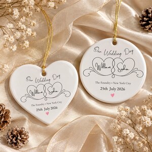May include: Two white ceramic ornaments, one heart-shaped and one circular, each featuring black text reading Our Wedding Day, William & Sophia, The Foundry, New York City, 25th July 2026, and a small pink heart, suspended by gold strings.