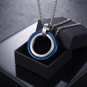May include: A silver-toned metal pendant featuring two interlocking rings, one with a smooth blue finish and the other with a brushed silver finish, suspended from a silver-toned box chain necklace resting on a small black jewellery box.