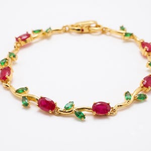 Op de afbeelding: Een goudkleurige armband met een rankachtige ketting met afwisselend ovaal geslepen rode stenen en marquise geslepen groene stenen. De metalen schakels zijn gevormd als stengels en bladeren die elke edelsteen in een cirkel verbinden.
