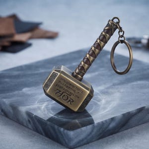 Könnte beinhalten: Ein bronzefarbener Metallschlüsselanhänger in Form eines Hammers mit strukturiertem Griff und einem runden Ring. Der Hammerkopf ist mit gravierten Knotenmustern und dem Text versehen: Whosoever holds this hammer, if he be worthy, shall possess the power of Thor.