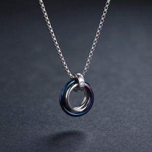 May include: A silver-toned box chain necklace featuring a pendant composed of three interlocking rings. The rings are finished in metallic blue, purple, and silver tones, with a small, clear, square-cut stone set into the top silver bail.
