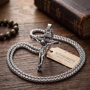 Puede incluir: Un colgante de crucifijo de metal plateado en una cadena trenzada gruesa. El colgante presenta una figura detallada de Jesús en la cruz con las letras INRI inscritas en la parte superior. Una pequeña etiqueta beige lleva impreso AETHELRED'S ARTIFACTS - EST. 2023.