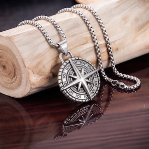 Peut inclure: Un collier pendentif en acier inoxydable argenté arborant un motif de boussole circulaire avec une étoile centrale proéminente. Le pendentif est suspendu à une chaîne carrée assortie, présentant des détails gravés et une finition métallique.