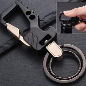 May include: A multi-functional keyring featuring a matte black carabiner body with metallic gold accents and a built-in bottle opener. The design includes a sturdy, dark gunmetal split ring for keys, showcasing a durable, industrial-style construction.