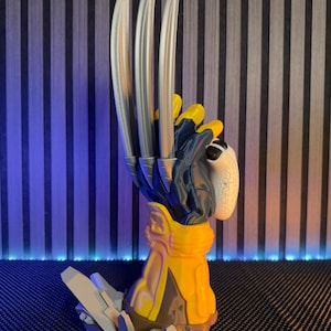 Pode incluir: Uma figura tridimensional de uma mão enluvada com três longas garras metálicas de cor prateada que se estendem para cima. A luva é principalmente azul-escura e amarela com uma palma branca texturizada. A base é uma estrutura rochosa cinzenta.