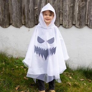 May include: A white hooded poncho featuring a dark grey graphic print of a jagged, toothy ghost face on the front. The garment is made of a lightweight, semi-sheer fabric with a pointed hood and a loose, flowing silhouette that drapes over the body.