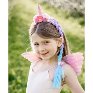 Peut inclure: Un serre-tête licorne avec une corne centrale, un groupe de fleurs en tissu rose et violet, et une longue extension de cheveux tressés bleus. Le serre-tête est doté de deux accessoires en forme d'ailes rose clair à texture matelassée.