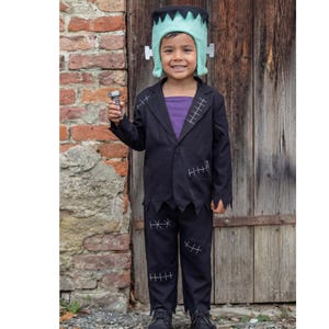 Peut inclure: Un costume inspiré de Frankenstein composé d'un blazer noir et d'un pantalon assorti avec des détails de coutures blanches. La tenue comprend une chemise violette en dessous et une coiffe vert clair à bord dentelé. Un boulon en plastique gris est tenu.