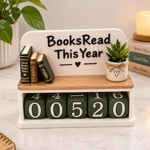 May include: A white wooden shelf with the text BooksRead This Year in black, featuring three miniature books, a small potted succulent in a white ceramic pot with a twine bow, and five green rotating number blocks displaying the digits 00520.