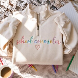 May include: A cream-coloured quarter-zip sweatshirt featuring the embroidered text "school counselor" in a multi-coloured script font. Below the text is a small, simple embroidered heart outline in a matching pink thread.